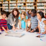 Teacher working with a diverse group of students reading and writing at a library table demonstrating how STEM education and literacy skills develop together through integrated learning