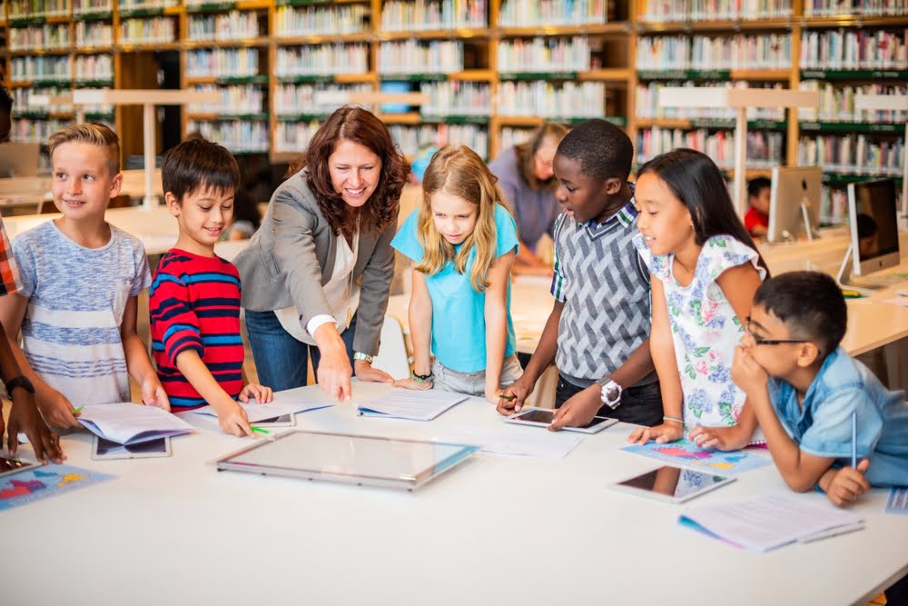 Teacher working with a diverse group of students reading and writing at a library table demonstrating how STEM education and literacy skills develop together through integrated learning